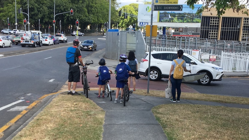 A parent with three schoolkids on bikes wait for a safe moment to cross a busy road