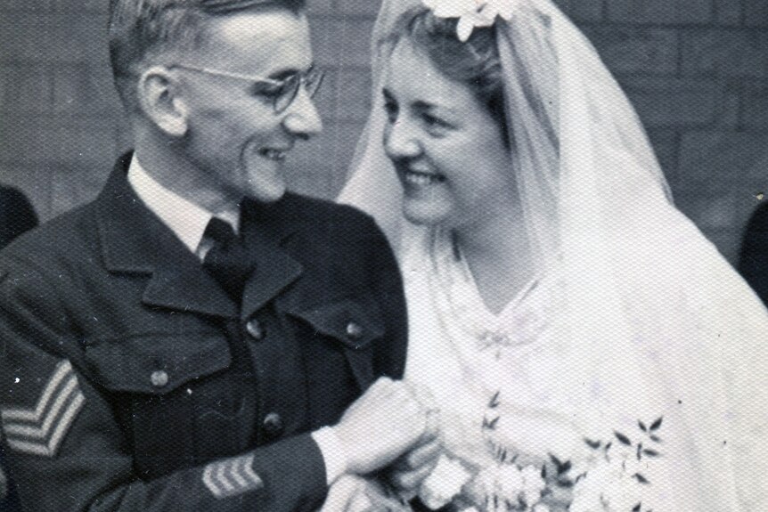 A black and white photo of a bride with a veil and flowers smiling at her groom in uniform