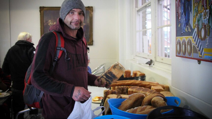 A man holding a load of bread, near a table full of bread.