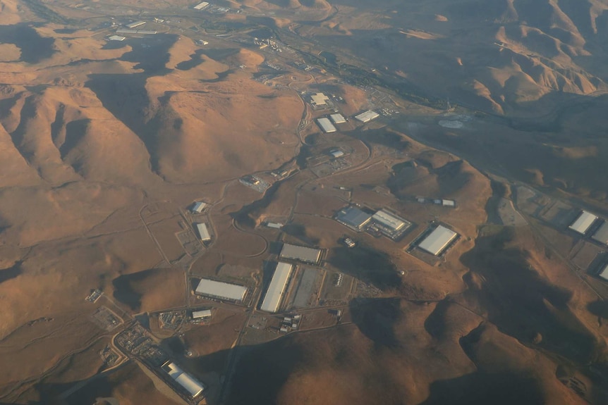 An aerial photograph shows brown desert peaks with silver industrial warehouses dotting a valley.