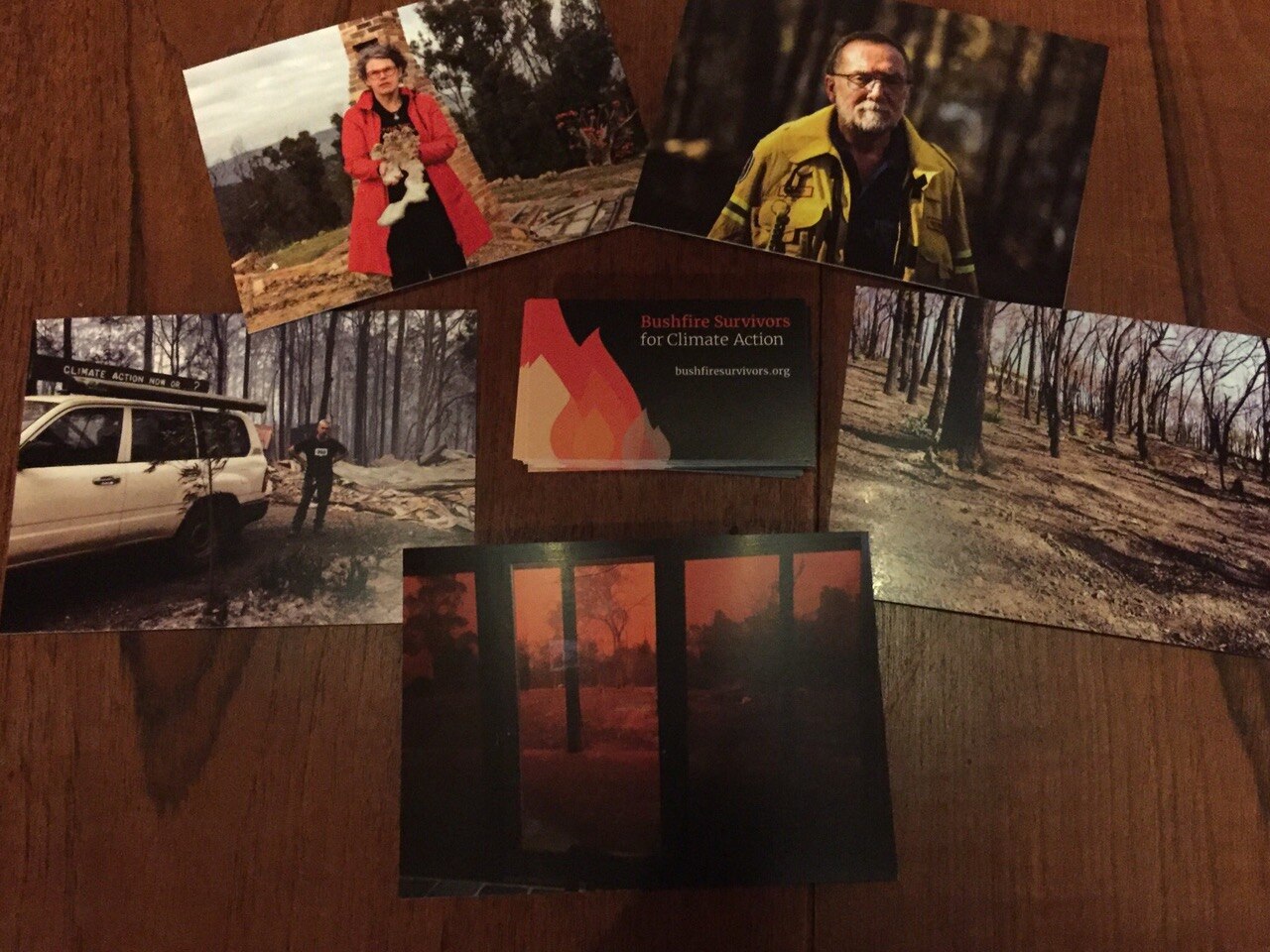 Postcards lay on a table: one shows a woman in front of a burnt home, and another shows a firefighter in burnt bushland.