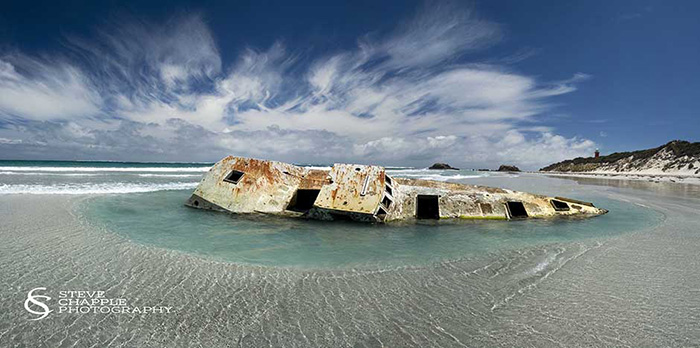 A rusted wrecked boat partially submerged in shallow water on a beach.