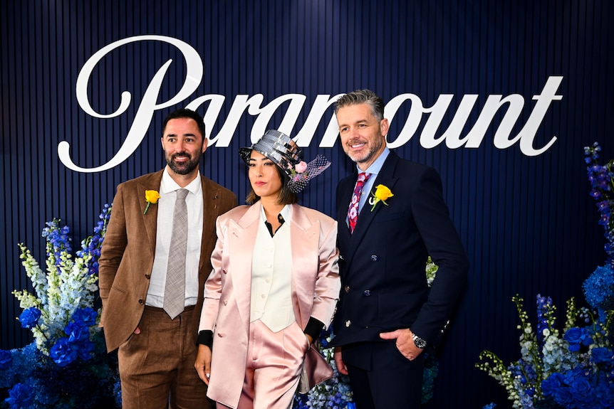 Two men and a woman in stylish clothes pose in front of a blue floral backdrop.