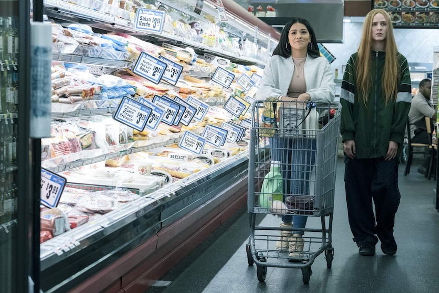 Gina Rodriguez pushing shopping trolley beside Evan Rachel Woods in brightly lit supermarket refrigerated aisle.