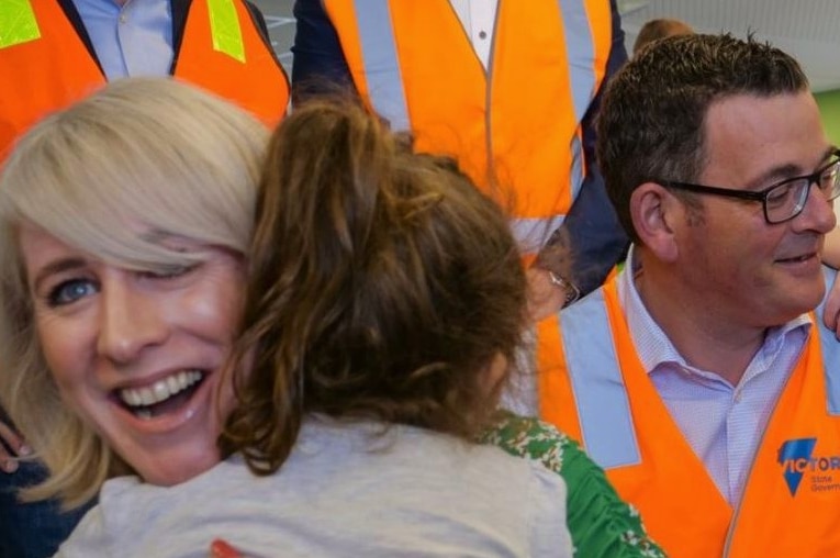 A woman with blond hair hugs a child and a man wearing glasses looks at a young girl.