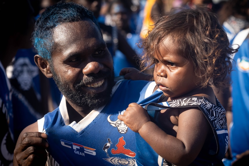 A young man holds his small child in his arms and smiles. Both are in identical jerseys.