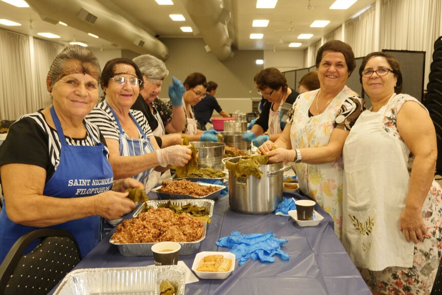 group of older Greek women smiling at camera around food preparation table.