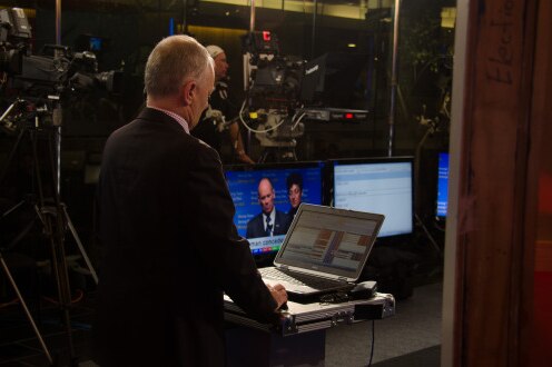 Antony Green stands in front of a computer screen in a busy news room