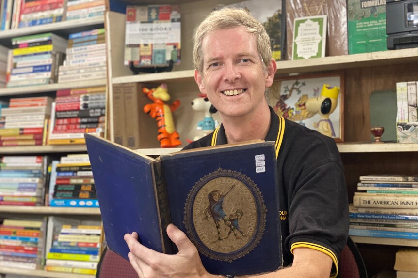 man holding a book, smiling a camera with book shelf behind him