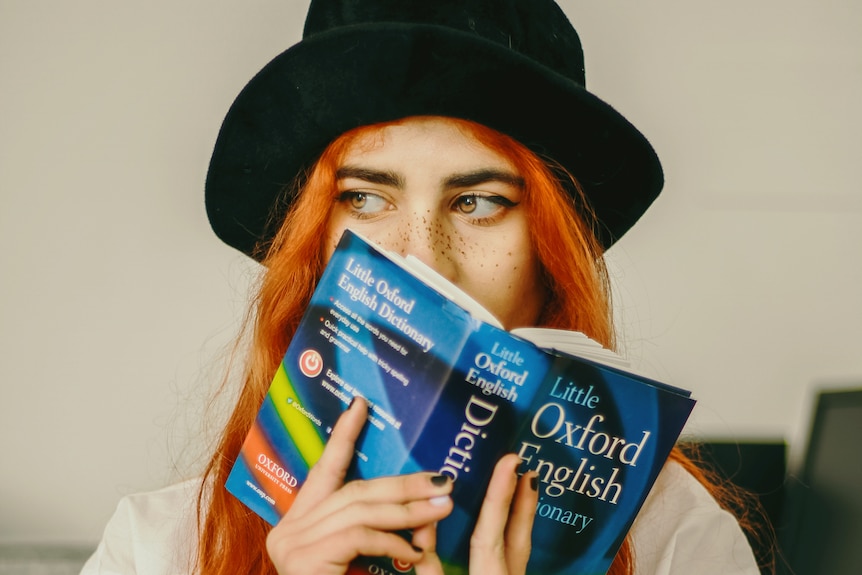 A woman with orange hair and a small black hat holds a small Oxford Dictionary close to her face. Only her eyes are visible.
