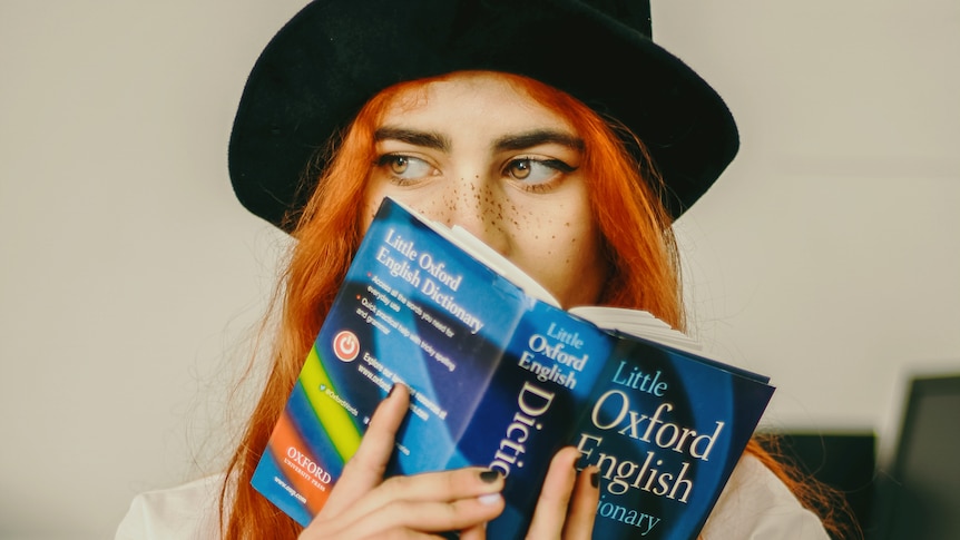 A woman with orange hair and a small black hat holds a small Oxford Dictionary close to her face. Only her eyes are visible.