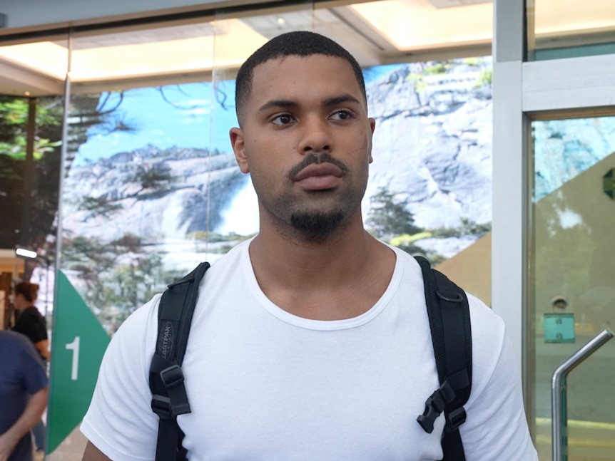 A dark-skinned man in a white T-shirt and black backpack.