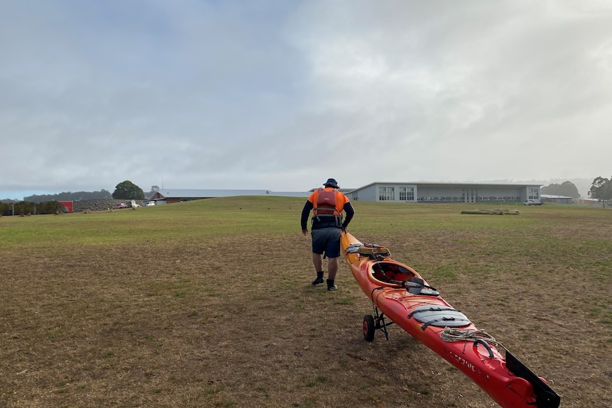 A man in a high-vis jacket walks up a hill towards a group of buildings carrying a kayak that is rolling on wheels 