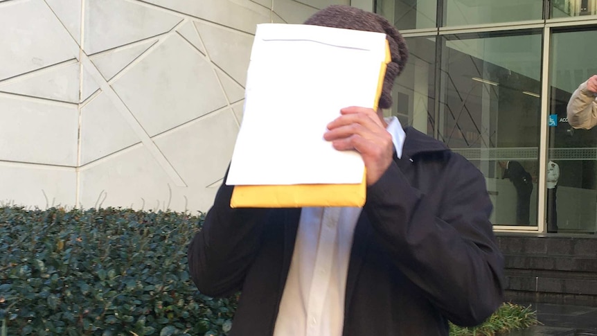 A mid shot of a man dressed in a white shirt and black jacket walking out of court holding up papers to cover his face.