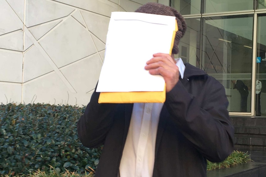 A mid shot of a man dressed in a white shirt and black jacket walking out of court holding up papers to cover his face.