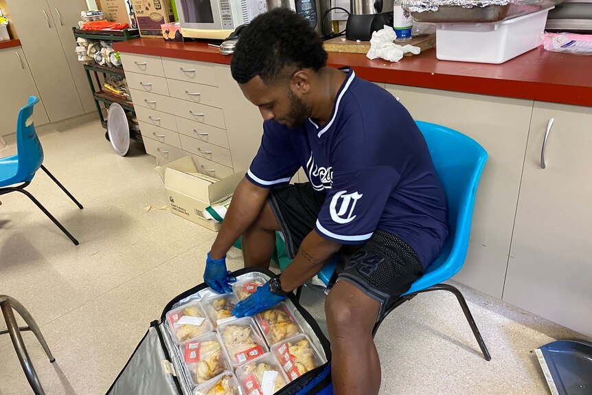 A man in a blue t-shirt sits on a chair and packs containers of food into a bag. 