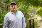 Matthew Hayden wearing a cap and grey jumper stands in a garden with trees behind him.