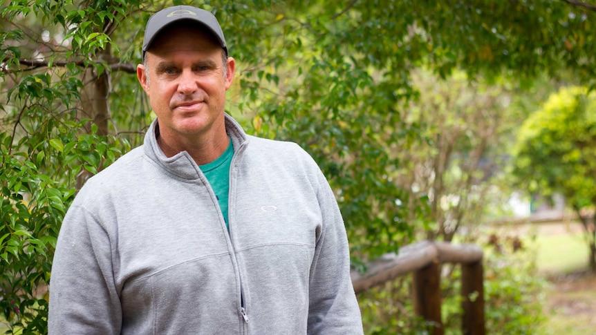 Matthew Hayden wearing a cap and grey jumper stands in a garden with trees behind him.