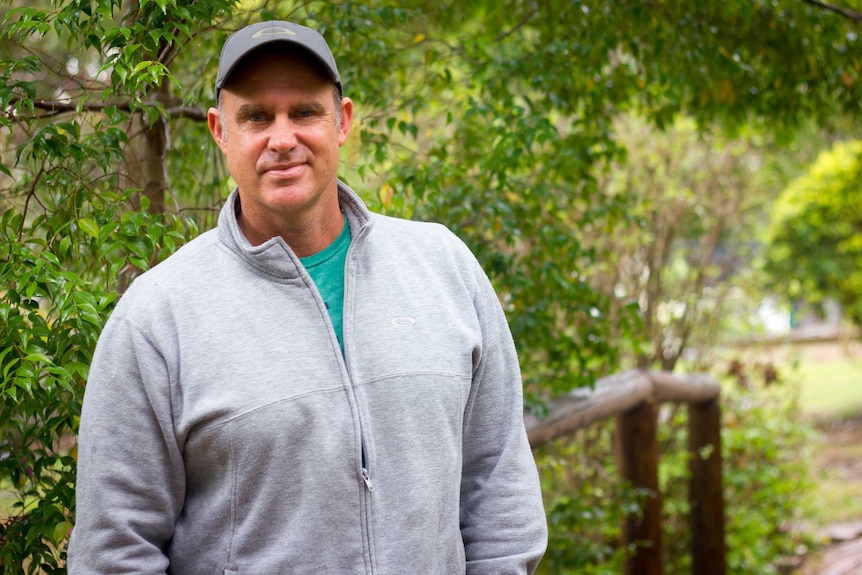 Matthew Hayden wearing a cap and grey jumper stands in a garden with trees behind him.