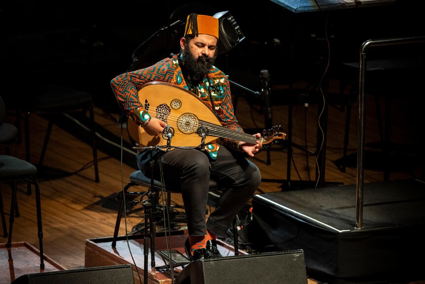 Joseph Tawadros sits on a chair on a cello riser on stage playing oud. He's wearing a red fez and brightly patterned shirt.