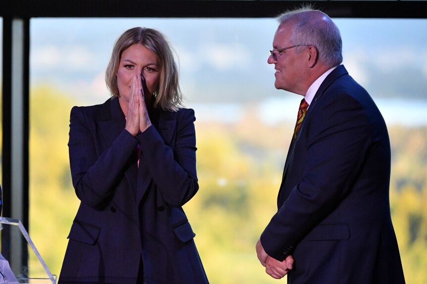 woman in navy blazer holds hand to face in shock as she stands on stage next to Prime Minster Scott Morrison