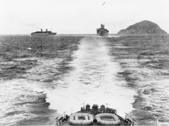 Two big dingys are on a boat deck and in the distance across the sea, two other war ships. It's a black and white image.
