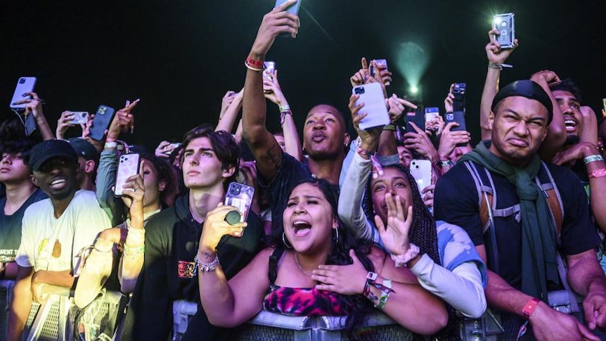 A mass of people scream and cheer from the front fence of a mosh pit holding mobile phones at night 