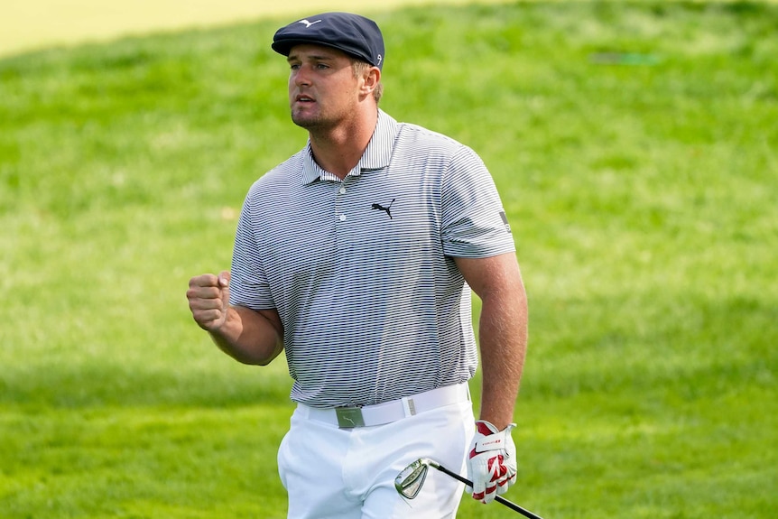 Bryson DeChambeau clenches his right fist and holds a club in his left, wearing a blue flat cap