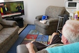 An elderly man with grey hair and glasses sits in his lounge room watching the news on TV.