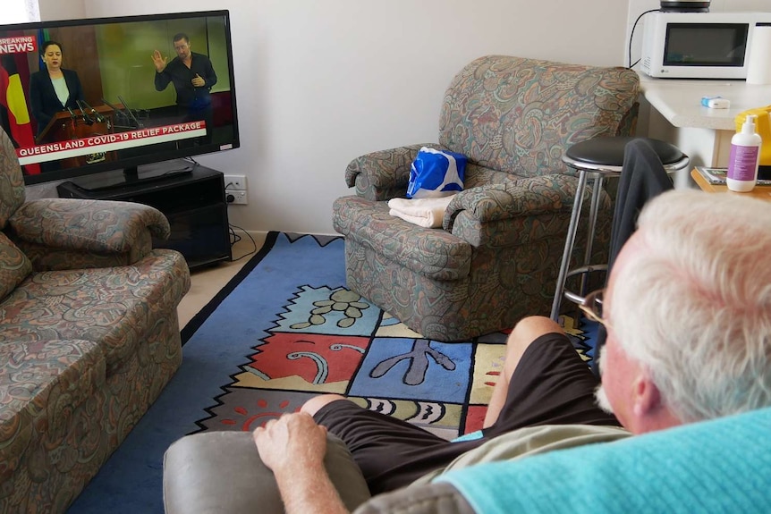 An elderly man with grey hair and glasses sits in his lounge room watching the news on TV.