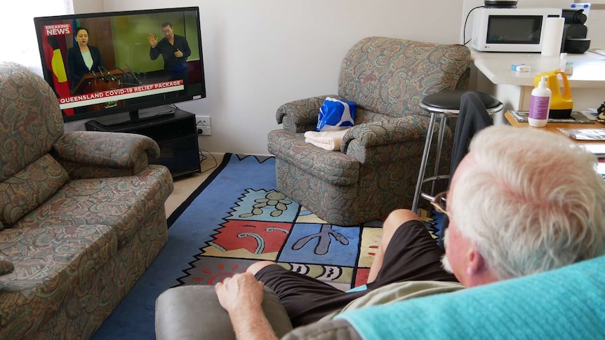 An elderly man with grey hair and glasses sits in his lounge room watching the news on TV.