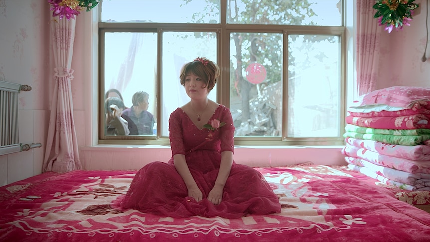 Colour still of Gai Qi seated and wearing a red wedding dress in a bedroom with people looking in through the window.