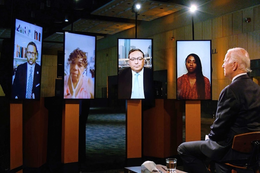 A man with white hair sits on a chair in a suit facing five screens with people.