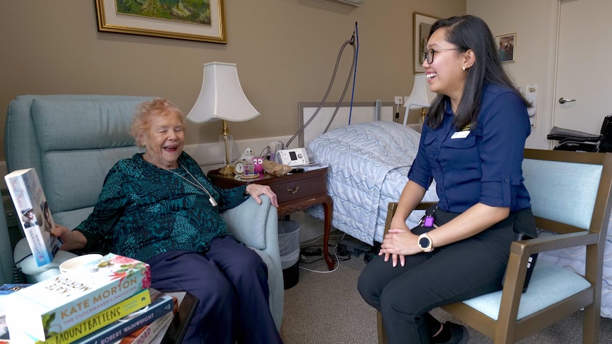 A 93-year-old woman and a younger nurse smiling and laughing in an aged care facility room.