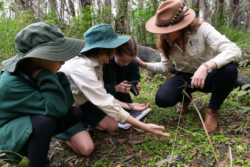 Three young students crouch down in the bush, taking a photo of flowers, with their principal