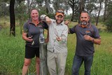 Three people holding bush tucker plants surrounded by scrub.