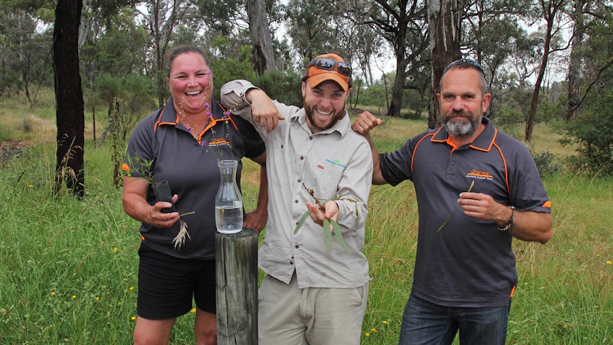 Three people holding bush tucker plants surrounded by scrub.