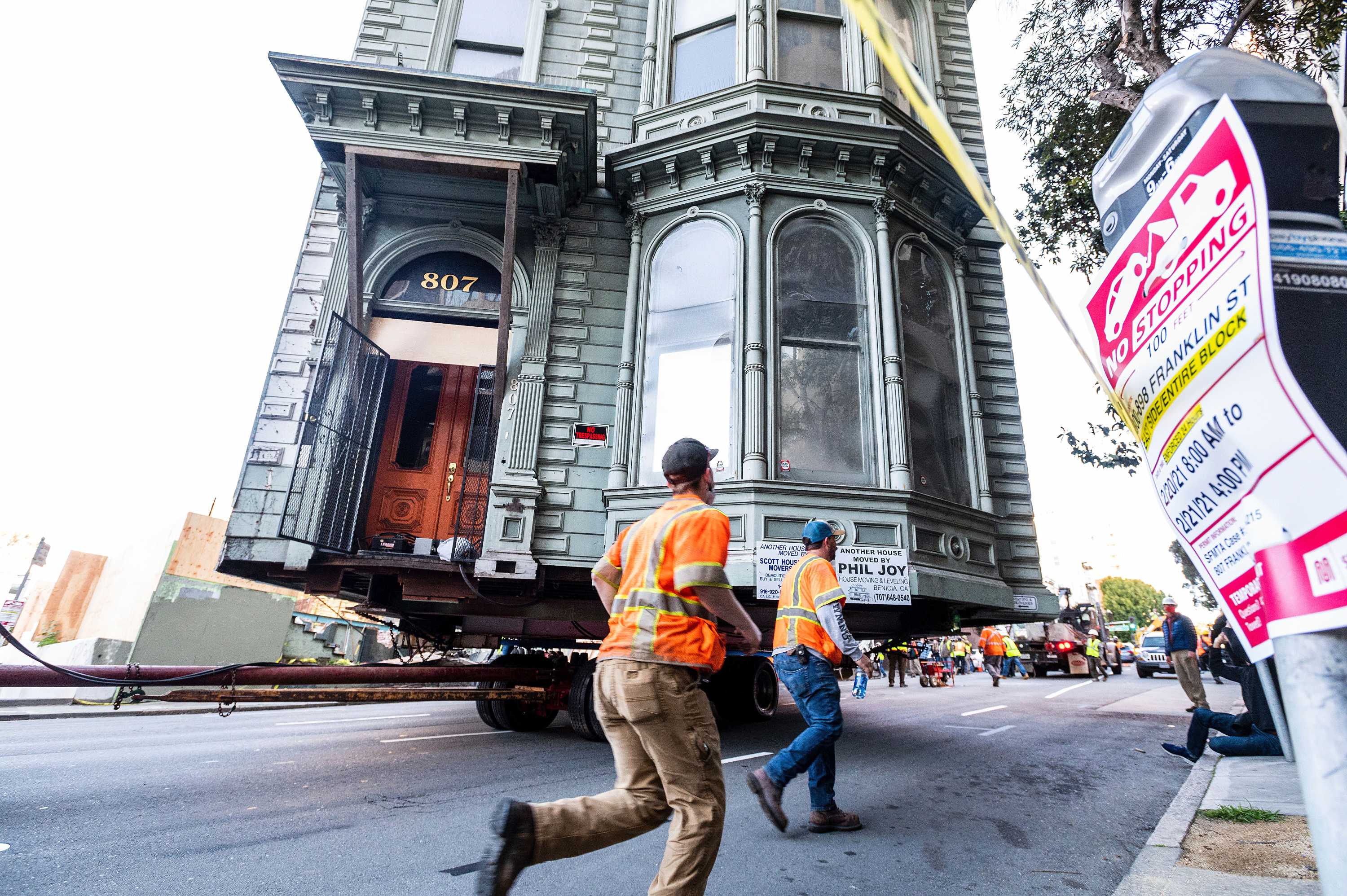 Two men are seen in front of a green home with a brown door and the number 807 on it as it's pulled through a street.