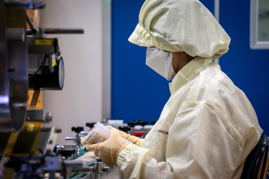 A woman wearing a mask with a white hair net and protective suit looks at her hands.