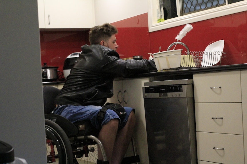 A young man in a wheelchair leans over his sink which is almost too high for him to access.