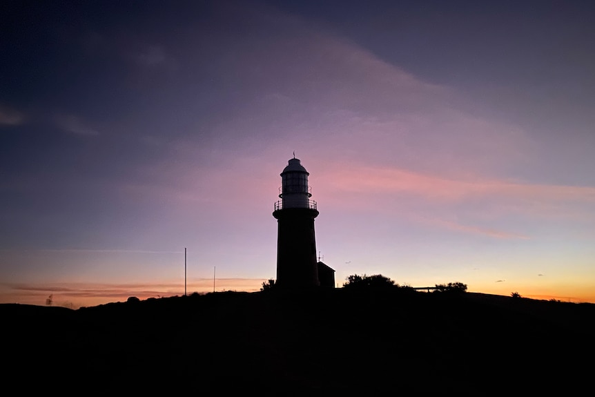 Black silhouette of a lighthouse is front of a purple-pink sunset with wisps of yellow. 