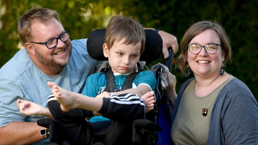 A young boy in a wheelchair with a smiling bearded man and a smiling woman, both wearing glasses