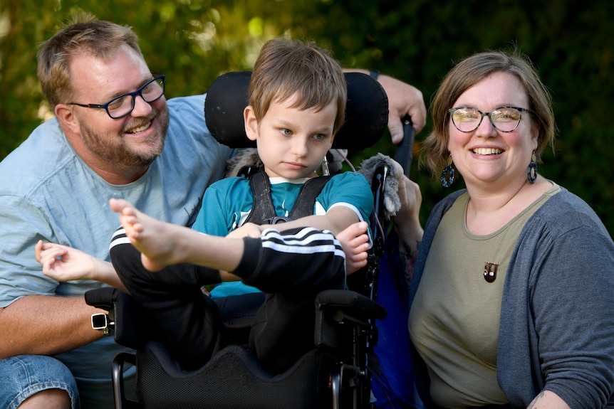 A young boy in a wheelchair with a smiling bearded man and a smiling woman, both wearing glasses