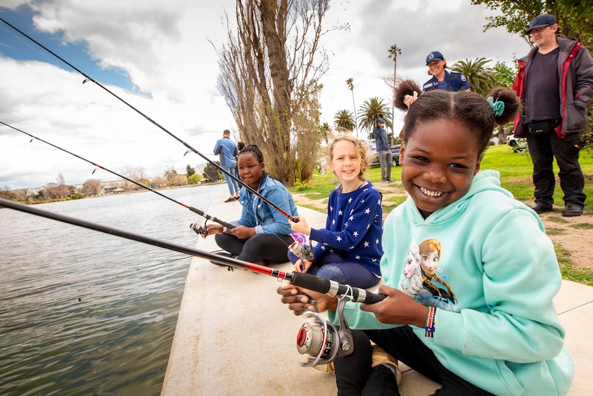 Three young girls smile at the camera while fishing on the bank of a river