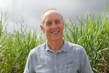 A man in a blue shirt smiles at the camera in front of a sugar cane field.