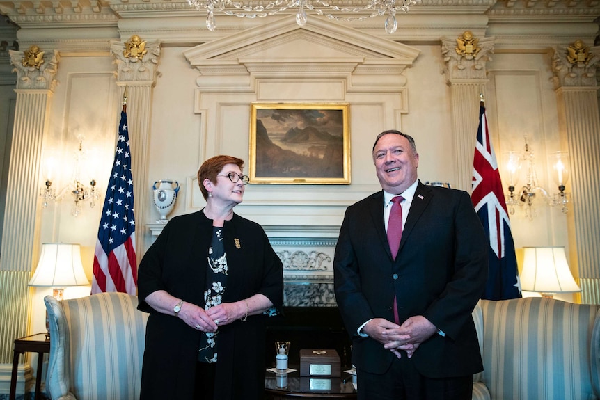 A woman with red hair and glasses stands net to a large man in a suit and tie in an opulent room with gold plated trim.