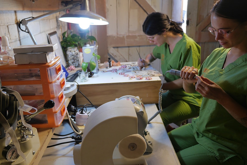Two women in green medical scrubs sit at a desk under a fluorescent light. One is painting the other uses a hand tool.