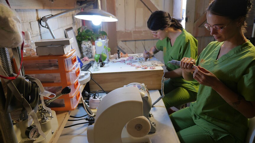 Two women in green medical scrubs sit at a desk under a fluorescent light. One is painting the other uses a hand tool.
