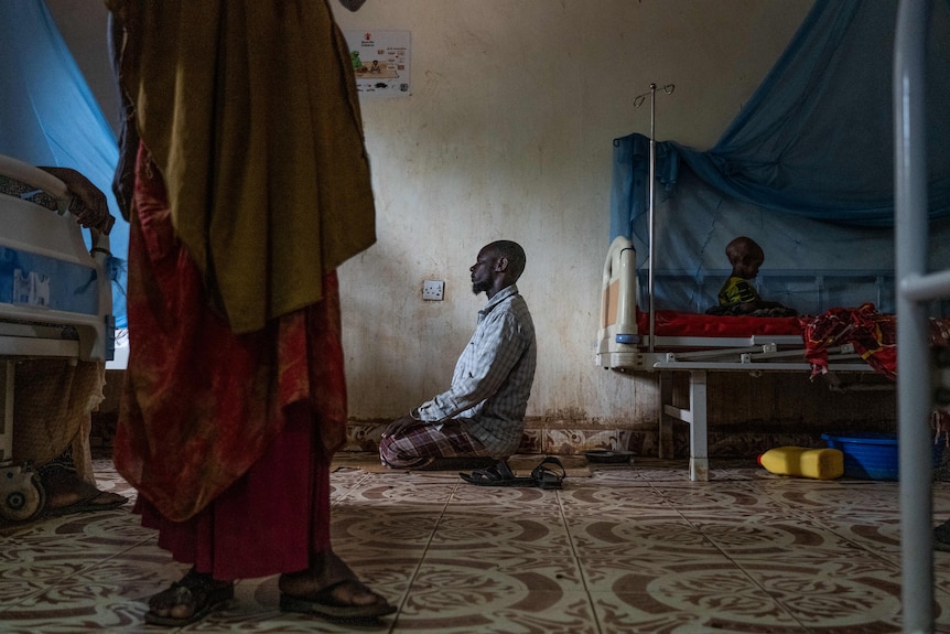 A man kneels on a prayer mat looking towards light from a window. Behind him a child sits on a hospital bed