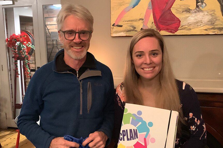 A man and a woman stand smiling next to each other, the woman holds a folder labelled 'cPLAN'.